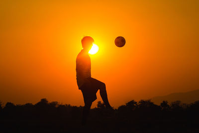 Silhouette man playing soccer on field against orange sky