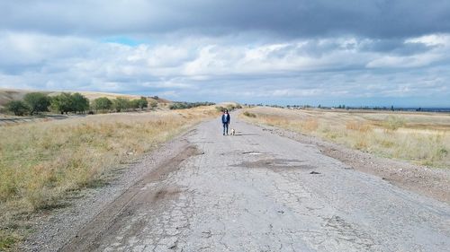 Distant view of woman standing with dog on road against cloudy sky