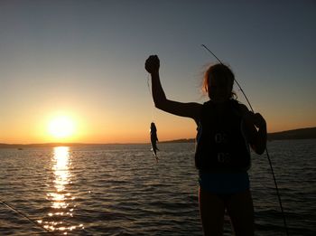 Girl holding dead fish in lake against sky during sunset