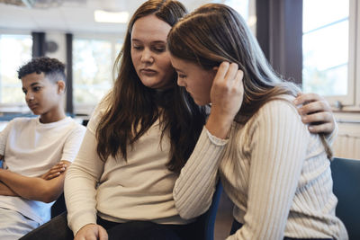 Sad teenage girl sitting with arm around female friend while consoling her in group therapy