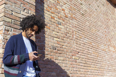 Young man using phone while standing against brick wall