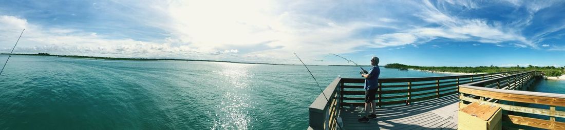 Panoramic image of man fishing while standing on pier by sea against cloudy sky