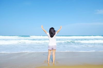Rear view of woman with arms outstretched standing at beach against blue sky