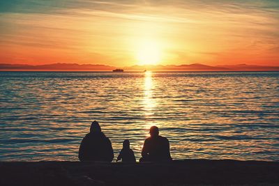 Silhouette people sitting by sea against orange sky during sunset