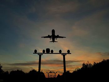 Low angle view of silhouette airplane flying over floodlights against sky during sunset