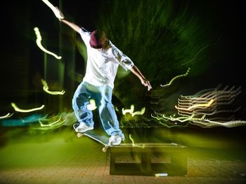 Man with arms outstretched skateboarding against light paints in park at night