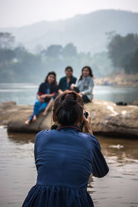 Rear view of woman photographing friends at lake against trees