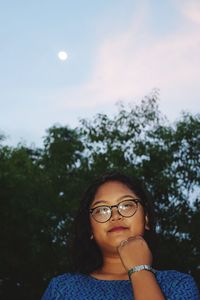 Low angle portrait of young woman wearing eyeglasses against trees during sunset