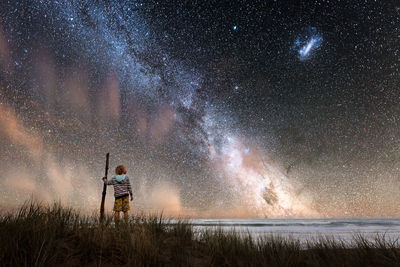 Boy holding walking stick standing at beach against starry sky