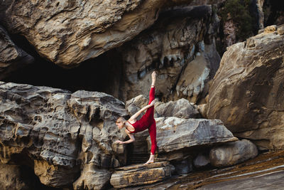 Side view of young woman in red sportswear doing downward facing dog pose standing on one leg while outstretching different leg on rough rock among mountains