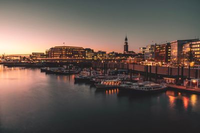 Boats moored in river against illuminated buildings in city at sunset