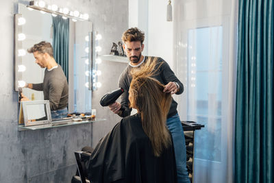 Ethnic male hairdresser drying hair of female client with closed eyes in modern beauty studio