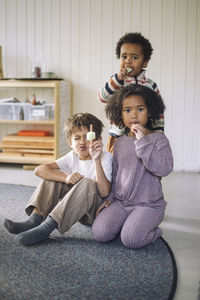 Portrait of boys and girl eating ice cream while sitting on carpet in classroom at kindergarten