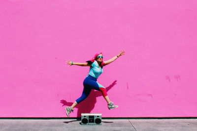 Full length of woman jumping over old-fashioned radio against pink wall