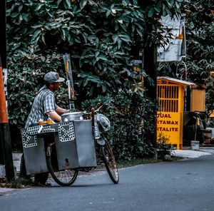 Side view of male vendor standing with bicycle on road against plants