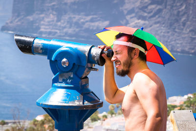 Side view of mid adult man with umbrella looking through coin-operated binoculars at beach