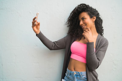 Portrait of young woman using phone while standing against wall