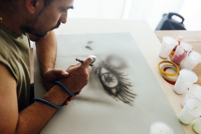 Aerial view of airbrush painter with cap drawing an eye on a paper.