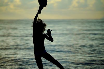 Silhouette boy with hand raised jumping by sea against sky during sunset