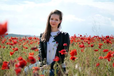 Portrait of young woman standing by poppy flowers on field against sky