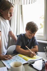 Smiling boy standing by brother doing homework at home