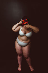 Young woman in lingerie holding mask standing against black background