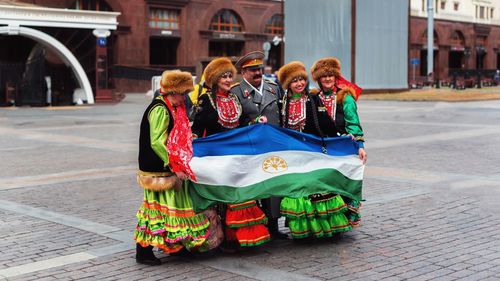 Women in traditional clothing and male army soldier holding bashkortostan flag on footpath