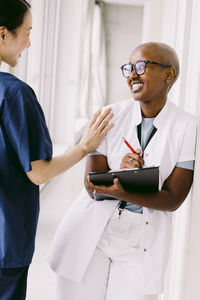 Happy female doctor and leaning with clipboard while looking at colleague in hospital corridor