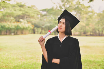 Thoughtful young woman in graduation gown holding certificate while standing at park