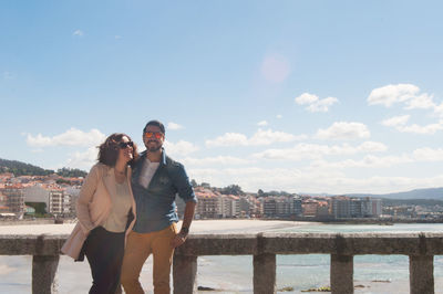 Smiling couple standing by railing at beach in city against sky during sunny day