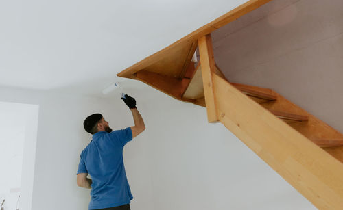 A young oriental man paints the ceiling under the stairs with a roller.