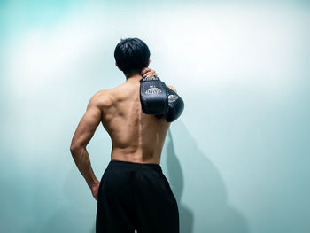 Rear view of shirtless mid adult man holding boxing gloves while standing against wall