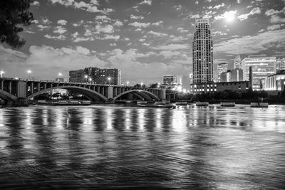 Illuminated bridge over river by buildings against sky at night