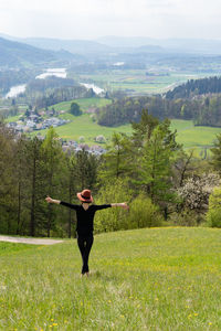 Young skinny woman with open arms and hat stands on a hill in nature of switzerland in springtime.