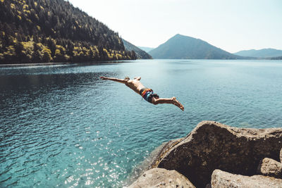 Side view of young male jumping in alpine lake in front of mountains
