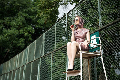 Full length of woman sitting on seat against fence