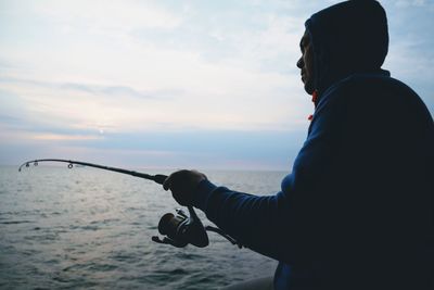 Side view of man fishing in sea against sky during sunset