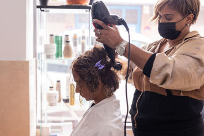 Side view of hairdresser wearing mask styling woman hair at salon