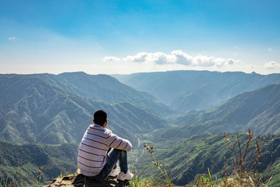 Rear view of man sitting on mountain against sky