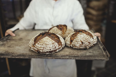 Midsection of baker holding fresh baked breads in tray at bakery