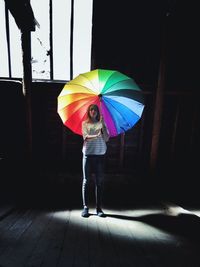 Woman holding colorful umbrella while standing by window in darkroom