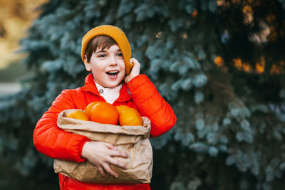 A boy in a bright orange jacket and a yellow hat holds a large package with oranges 