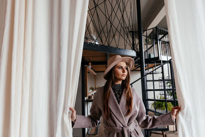 A girl in a beige hat and a beige coat stands indoors and pushes the white curtain with her hands