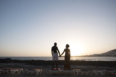 Couple standing at beach against sky during sunset