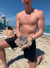 Full length of shirtless man holding sunglasses at beach against sky