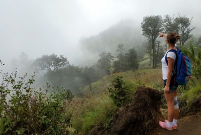 Full length of female hiker pointing on trees in forest during foggy weather