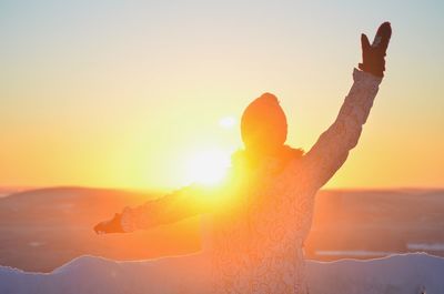 Rear view of woman with hand raised standing against sky during sunset