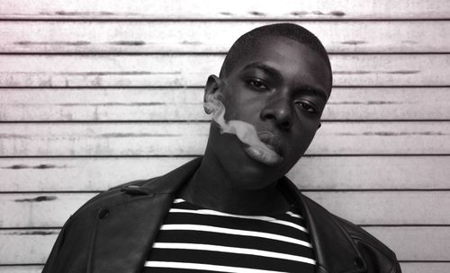 Portrait of young man exhaling smoke standing against corrugated iron