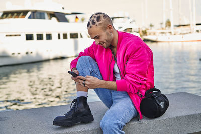 Positive eccentric male with stylish haircut in modern outfit browsing cellphone on marina with moored boats