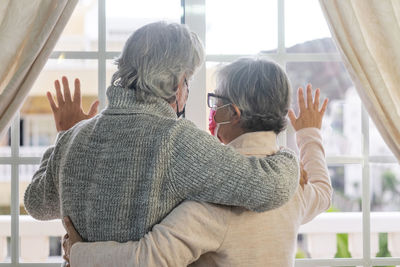 Rear view of senior couple wearing mask looking through window at home